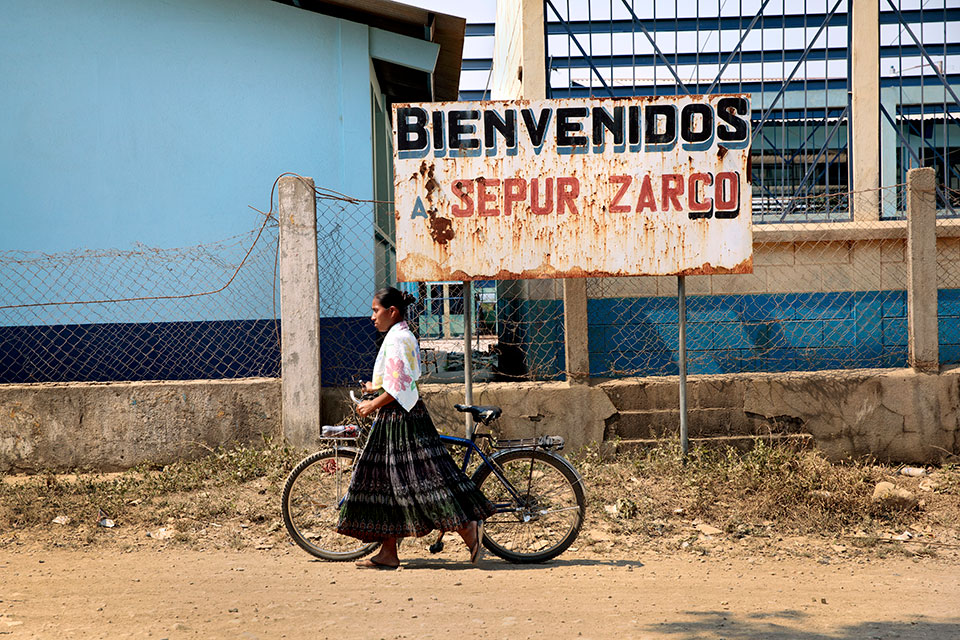 An indigenous woman walks a bicycle past a sign that reads "Welcome to Sepur Zarco" in Guatemala.