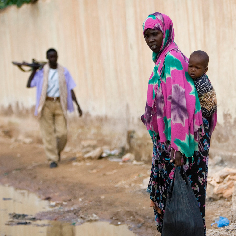 A woman carries a baby on her back. A man with a gun walks behind her.